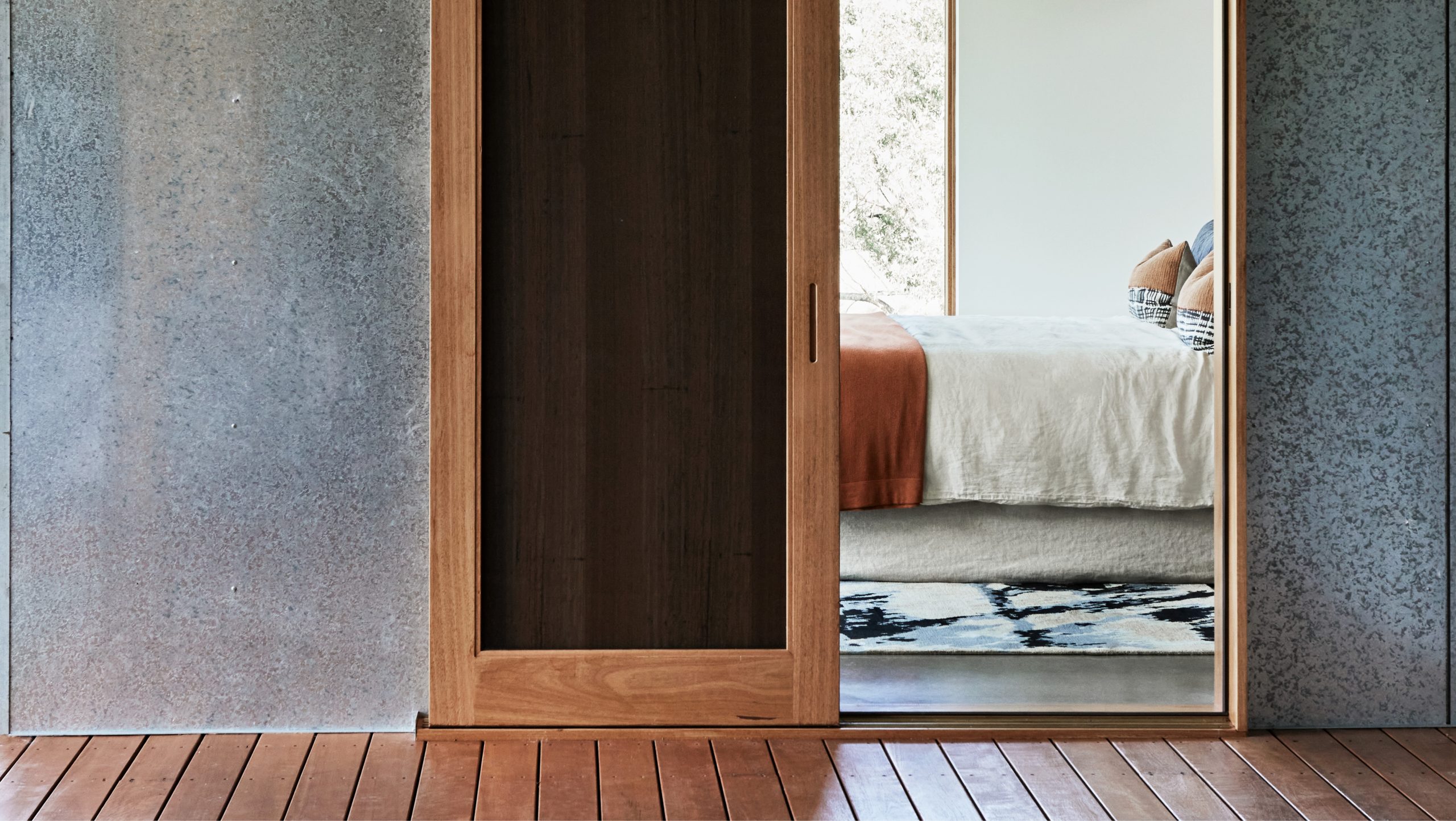 Bedroom reflected through a timber sliding door with concrete-panelled walls, Shores rug from the Hare + Klein x Designer Rugs fourth collection at the foot of the bed