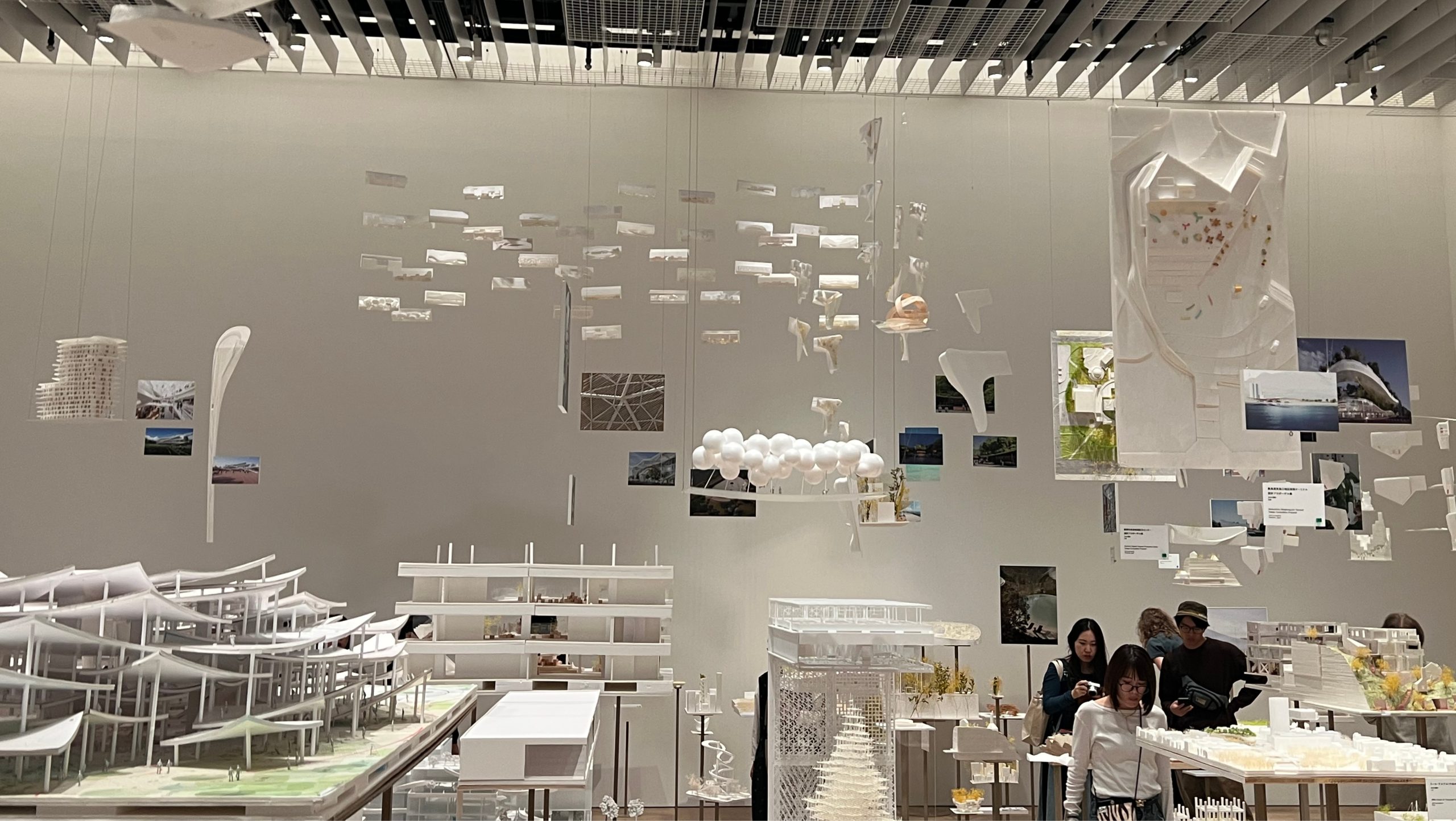A wide view of the Sou Fujimoto Mori Art Museum exhibition space in Tokyo, with white architectural models on tables, suspended sketches and photographs, and visitors viewing the display