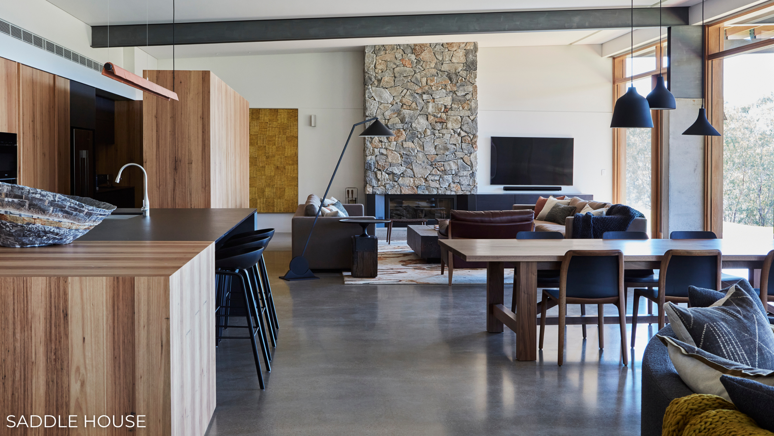 Saddle House living, kitchen and dining area by Cristina Repeti for Hare + Klein, with timber cabinetry, a stone fireplace and polished concrete floors